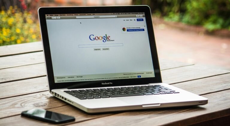 A MacBook Pro displaying Google Search on a wooden table outdoors, next to a smartphone.