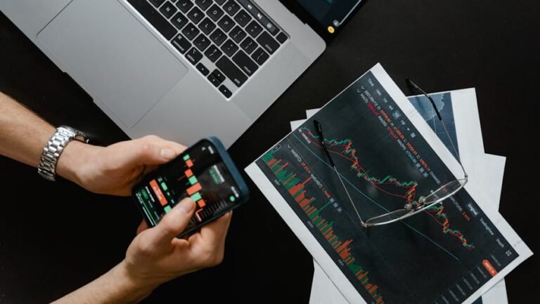 Hands using smartphone beside laptop with stock charts, showcasing digital trading.