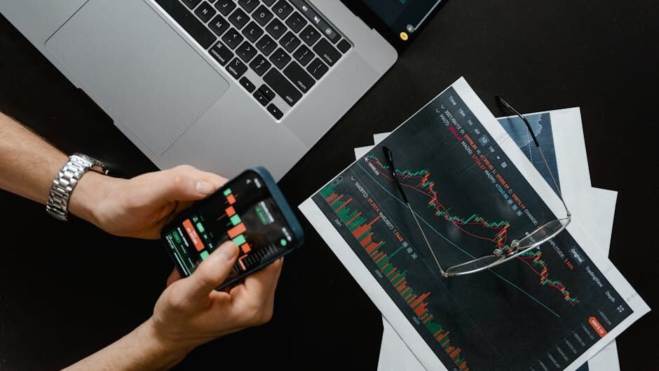 Hands using smartphone beside laptop with stock charts, showcasing digital trading.